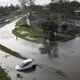 An aerial shot of a winding road, and house flooded by hurricane waters. A partially submerged car can be seen on the road.